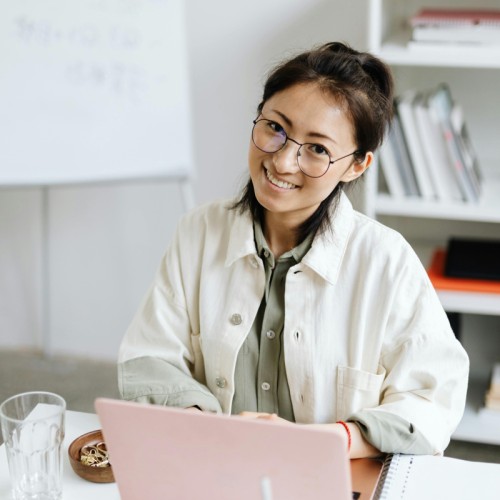 A person with glasses sat a desk, smiling at the camera behind a pink laptop